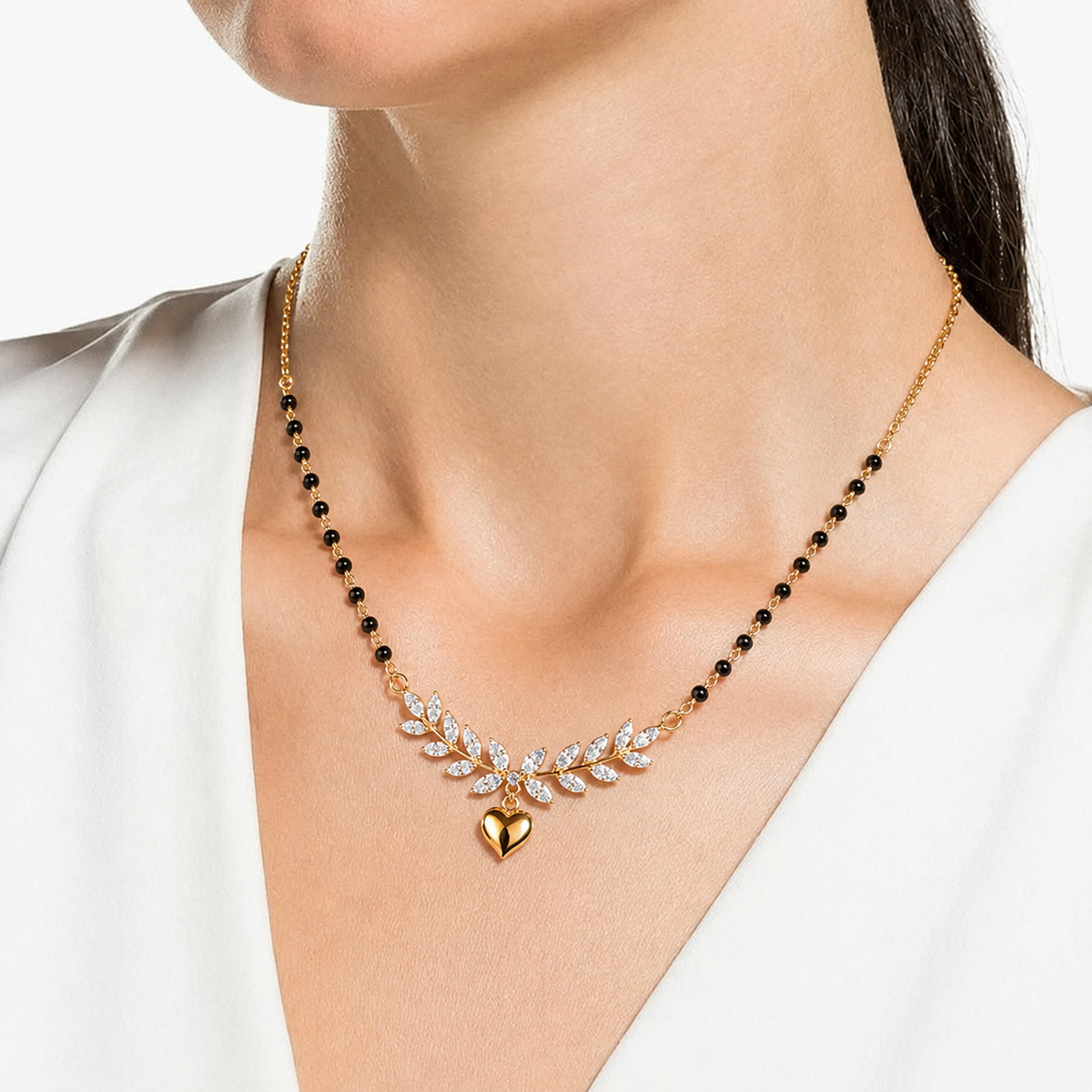 A close-up of a model wearing a delicate gold necklace with black beads, a leafy crystal pendant, and a small gold heart charm.